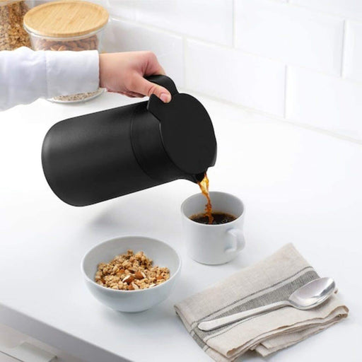 A stainless steel vacuum flask with a black lid and handle, filled with hot coffee, sitting on a wooden table. 50360231