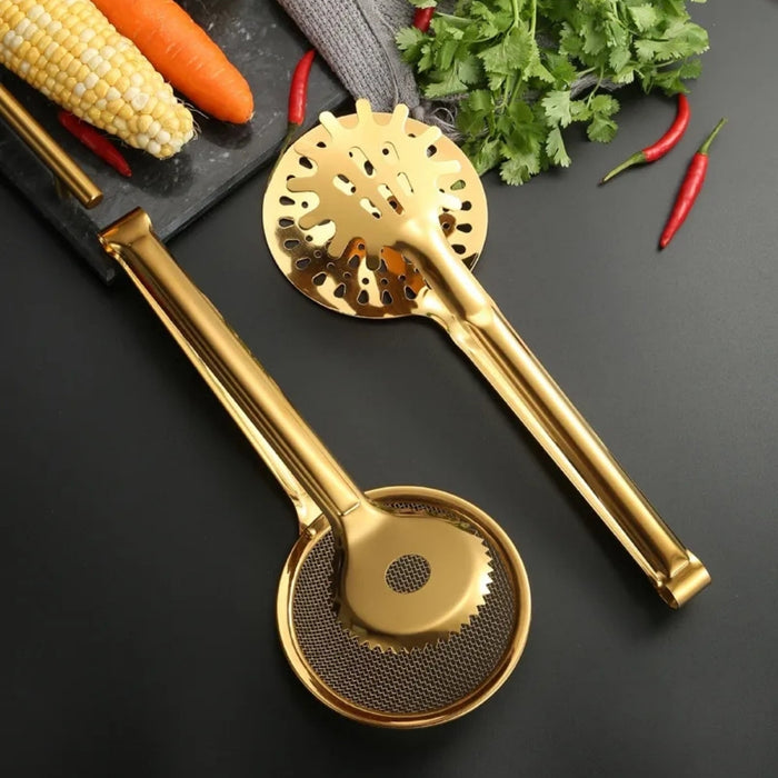 Two golden stainless steel kitchen tools—a mesh strainer tong and a slotted skimmer tong—resting on a dark backdrop. One features a circular wire mesh head with serrated edges, while the other has a starburst perforated design with round holes. Nearby are fresh ingredients: corn cob, carrots, red chilies, and cilantro, arranged on a matte black kitchen tray.- 1600003112384 13034149
