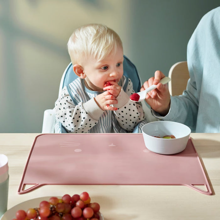Child seated in a high chair eating fruit with an adult’s assistance, using a pink cat-face silicone placemat on the table.

