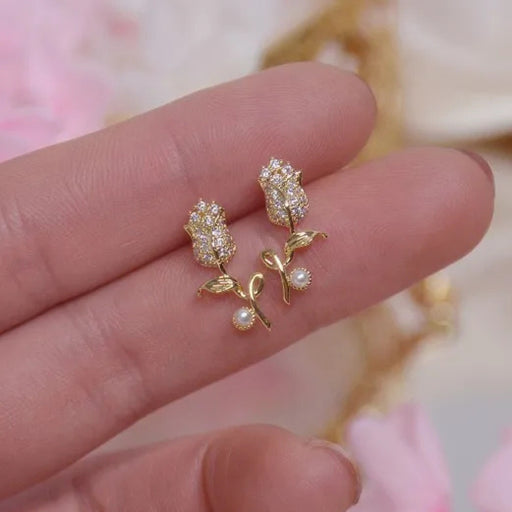 Gold rose-shaped earrings with pearls held between fingers against a blurred floral background