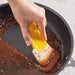 Hand using yellow silicone scraper to clean reddish-brown residue from non-stick pan – 1598449174494 14075321
