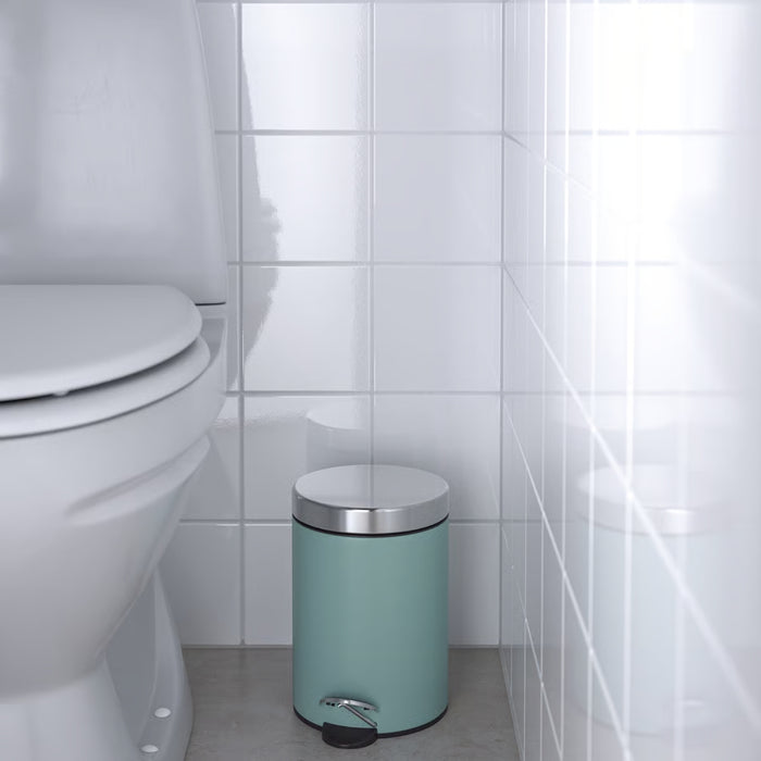 Teal trash bin with silver lid next to a toilet in a bathroom.