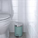 Teal trash bin with silver lid next to a toilet in a bathroom.
