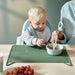 Child reaching for fruit at a table with a green silicone placemat, assisted by an adult holding a spoon.

