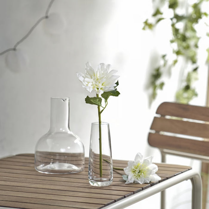 Clear vase with a single white flower on a wooden table outdoors.