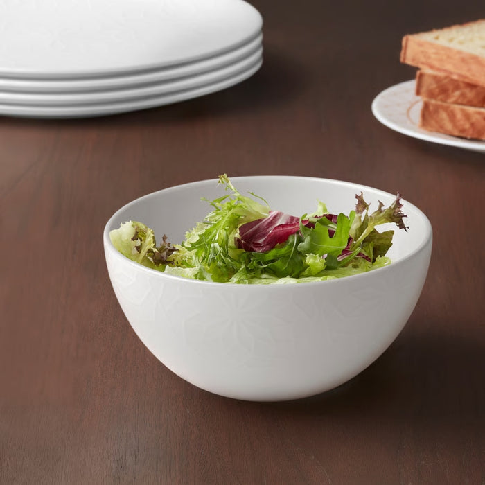 White bowl of salad on a wooden table with plates and bread in the background