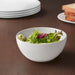 White bowl of salad on a wooden table with plates and bread in the background
