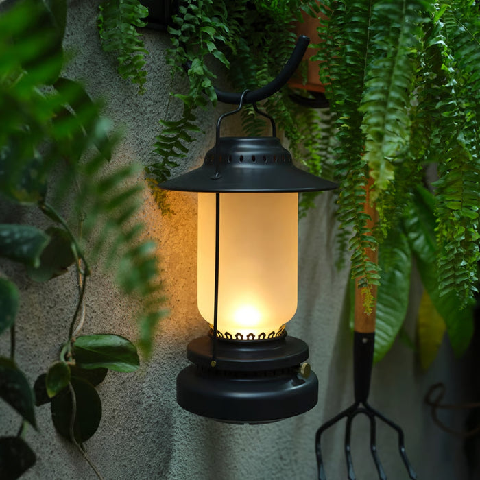Black lantern with warm glow surrounded by green plants against a textured wall.