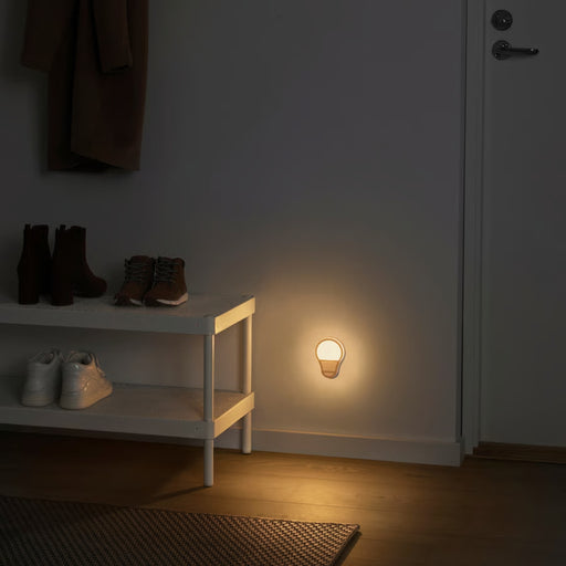 Warmly lit bulb-shaped nightlight plugged into a wall socket in a hallway, softly illuminating a shoe rack with boots and sneakers, a hanging coat, and a wooden floor with a textured mat.