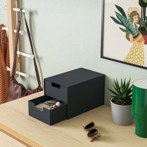 Black storage box with a drawer on a wooden surface, surrounded by a plant, green cup, and sunglasses.