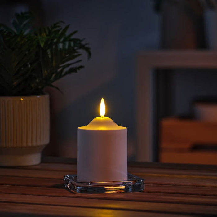 SMÅLOM candle lit on glass holder, emitting warm glow beside potted plant in cozy indoor setup