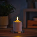 SMÅLOM candle lit on glass holder, emitting warm glow beside potted plant in cozy indoor setup