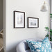 Modern interior corner with gray armchair, blue floral pillow, and two framed photos on white wall - 70327189