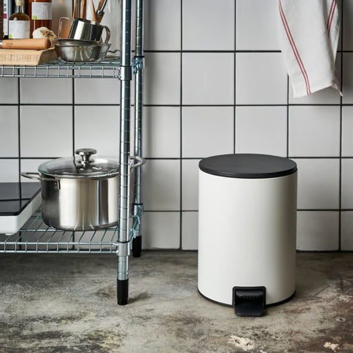 White pedal bin placed in a kitchen setting next to a metal wire shelf unit.