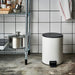 White pedal bin placed in a kitchen setting next to a metal wire shelf unit.