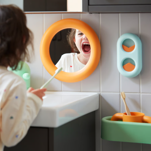 Child holding a toothbrush at a bathroom sink, looking into a round orange-framed mirror with mouth open.
