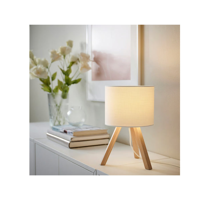 Table lamp with wooden base and white shade on a surface with books and flowers in the background.