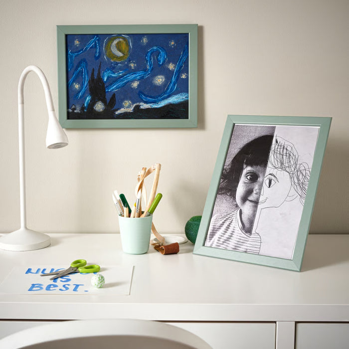 IKEA FISKBO frame in light grey-green, 21x30 cm (8 ¼x11 ¾") placed on desk beside “HUGS IS BEST” note and stationery; displays child’s drawing in playful workspace with Starry Night wall art and white lamp – ideal for creative home decor - 10584916