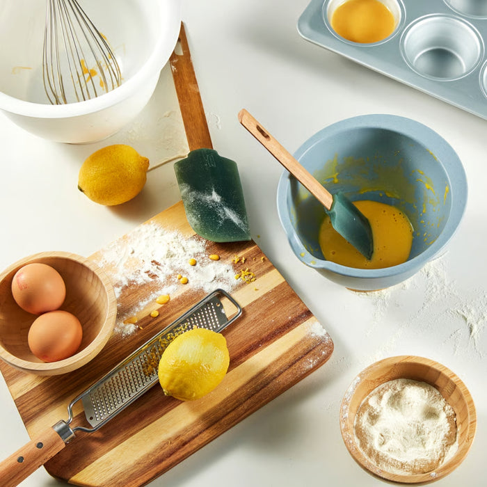 A kitchen scene with baking ingredients and utensils, including a white mixing bowl with a whisk, a lemon, a green spatula, a wooden cutting board with flour and lemon zest, a metal grater, a wooden bowl with eggs, a blue mixing bowl with a yellow mixture, and a muffin tin with one filled cavity-90607438