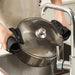 Person using a black Multi-Functional Silicone Anti-Skid Heat Insulation Clip Guard to securely grip the lid of a stainless steel pot while draining hot water into a sink. Steam rises visibly as the pot tilts, with a chrome faucet and tiled backsplash in the background—highlighting the tool’s heat resistance and anti-slip function during everyday kitchen tasks -1598452898401 13017586.
