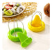 Two plastic fruit slicer/corer tools on a white surface, one green and one yellow, with metal wire loops and triangular cutting pieces. A plate of sliced fruit and a kiwi appear blurred in the background as props.