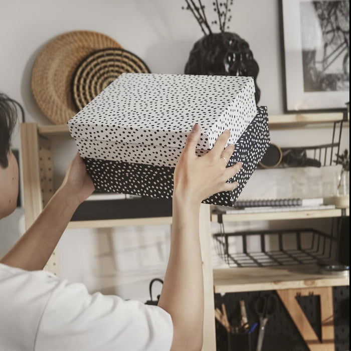 IKEA RÄTTAREN storage box set—person placing two polka dot boxes onto a wooden shelf; top box is white with black dots; bottom box is black with white dots. Surrounding shelf items include a woven basket, black vase with branches, framed photo, and notebooks, creating a cozy and organized home-office vibe – 90597986.