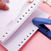 Stack of white loose-leaf papers with evenly spaced round holes along the edge, secured by silver binder rings. A blue metal hole puncher lies beside the papers, showcasing its role in creating the aligned holes. The desktop setup reflects a neat and organized approach to document handling—ideal for office, school, or personal filing systems – 1598448803339 13141950.