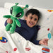 Child cuddling a green alien plush toy with antennae and big eyes, lying in space-themed bedding decorated with planets and stars, creating a joyful bedtime atmosphere.