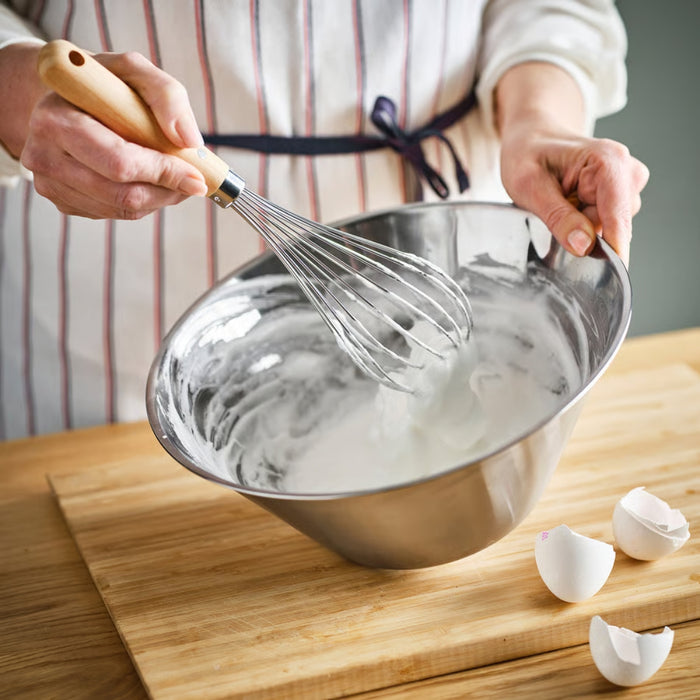 A person is whisking a creamy white mixture—likely egg whites or batter—in a stainless steel mixing bowl using the IKEA VARDAGEN balloon whisk. The individual is wearing a white apron with red vertical stripes and a navy waist tie. The setup is placed on a wooden countertop with two cracked eggshells beside the bowl, emphasizing a natural kitchen scene of hands-on food preparation -00581485.