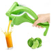 A green manual citrus juicer being squeezed by a hand, with orange juice splashing into a glass and limes shown below on a white background.