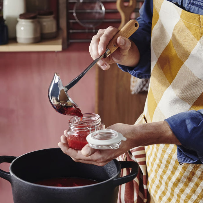 Person in a kitchen pouring jam into a jar with a ladle.