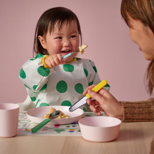 Child seated at table wearing white shirt with green polka dots; adult assists with IKEA SMASKA yellow-handled knife from 3-piece cutlery set – pink bowls and cup on illustrated placemat create playful mealtime scene - 70596940