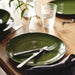 Close-up of a green PELARKAKTUS side plate with a silver fork and black-handled knife on a wooden dining table.