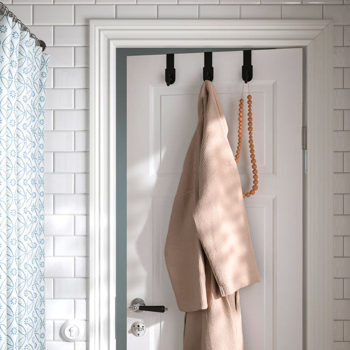 IKEA VALASJÖN hanger in use on a white bathroom door, holding a beige bathrobe and wooden bead decor beside a patterned shower curtain