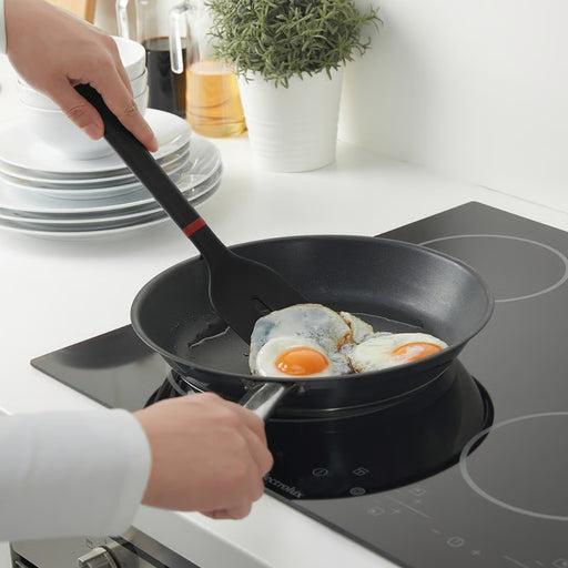 A person uses a black spatula with a red accent to lift a sunny-side-up egg in a black frying pan on a modern induction cooktop, with white plates, a potted plant, and cooking oil in the background.