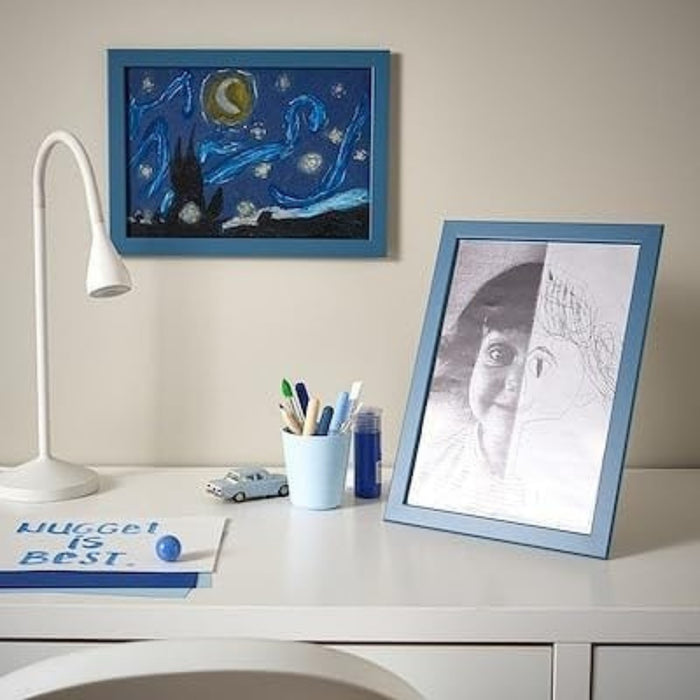 IKEA FISKBO frame in dark grey-blue, 21x30 cm (8 ¼x11 ¾") placed on desk; displays split portrait of child’s photo and drawing – styled with white lamp, pen holder, toy car, and “NUGGET IS BEST” note for playful workspace decor - 20584930