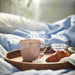 IKEA FÄRGKLAR Mug, light pink, 42 cl, placed on a wooden tray alongside a croissant, jam jar, and a glass of water, set on a bed with blue sheets. The smooth matte finish and simple, curved shape enhance its modern appeal.-90604883