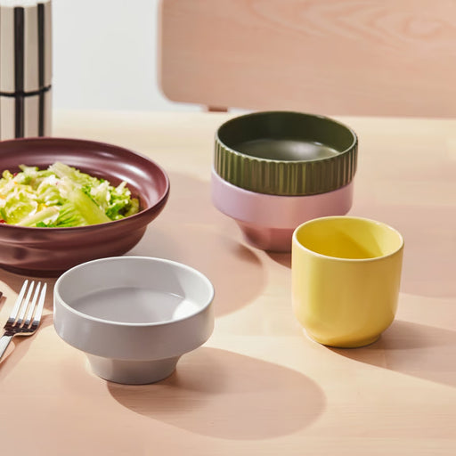 SPJUTLILJA bowls styled on dining table with maroon salad bowl, white wide-rim bowl, yellow cup, and stacked green ribbed and pink bowls