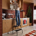 Person using BÄSINGEN 2-step stool with handrail in anthracite to reach upper kitchen cabinets. Set in a warm-toned kitchen with red tiles, wooden cabinetry, and a cactus plant.– 30595754