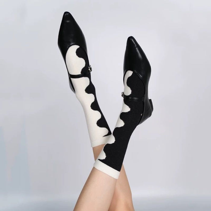 Legs raised wearing black pointed shoes and mismatched wave socks against gray background