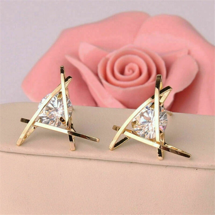 Triangle gold earrings – round crystal in intersecting gold triangle frame, placed on soft fabric with pink rose backdrop

