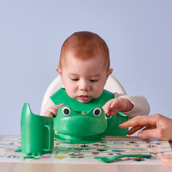 Baby wearing a green frog-shaped bib at a table with a plain background