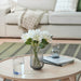 Round coffee table with a vase of white flowers, two glasses, and a book in a living room setting.