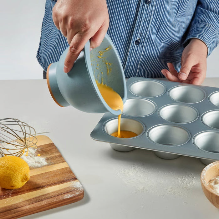 A person wearing a blue and white striped shirt pouring batter from a mixing bowl into a muffin tin. The wooden cutting board holds a lemon, flour, and a whisk, with scattered flour adding a natural baking touch.-90607438