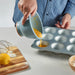 A person wearing a blue and white striped shirt pouring batter from a mixing bowl into a muffin tin. The wooden cutting board holds a lemon, flour, and a whisk, with scattered flour adding a natural baking touch.-90607438