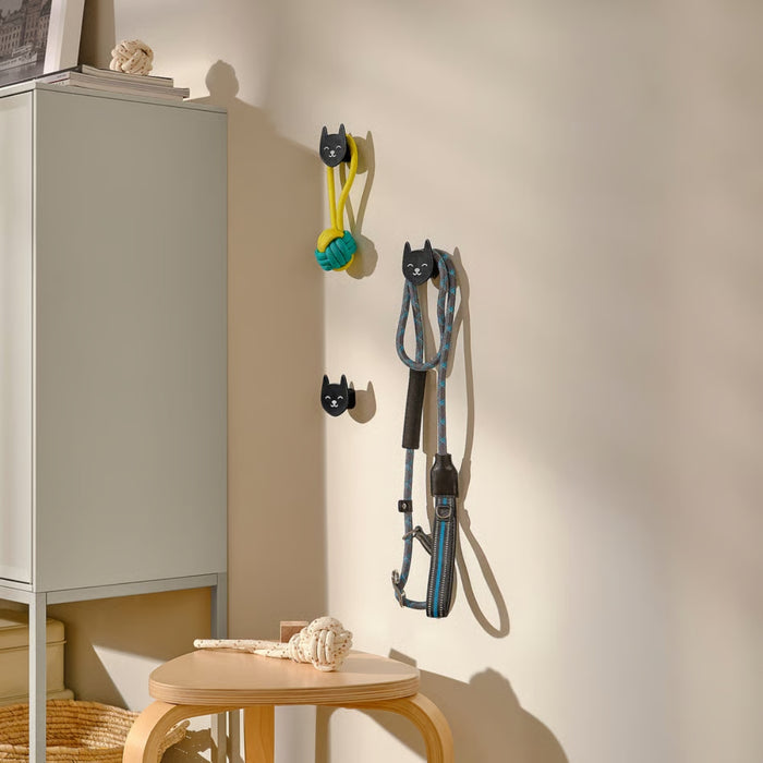 Three black cat-shaped wall hooks mounted on a light-colored wall, holding a yellow and green rope toy, a blue and black leash, and one empty. Below them, a small wooden stool holds a white rope toy, creating a stylish and practical pet accessory storage area.-90567791