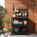 A well-organized outdoor gardening station with a black metal shelving unit against a brick wall. The shelves hold woven baskets in green, blue, and white filled with gardening tools, spray bottles, and terracotta pots.-10591422