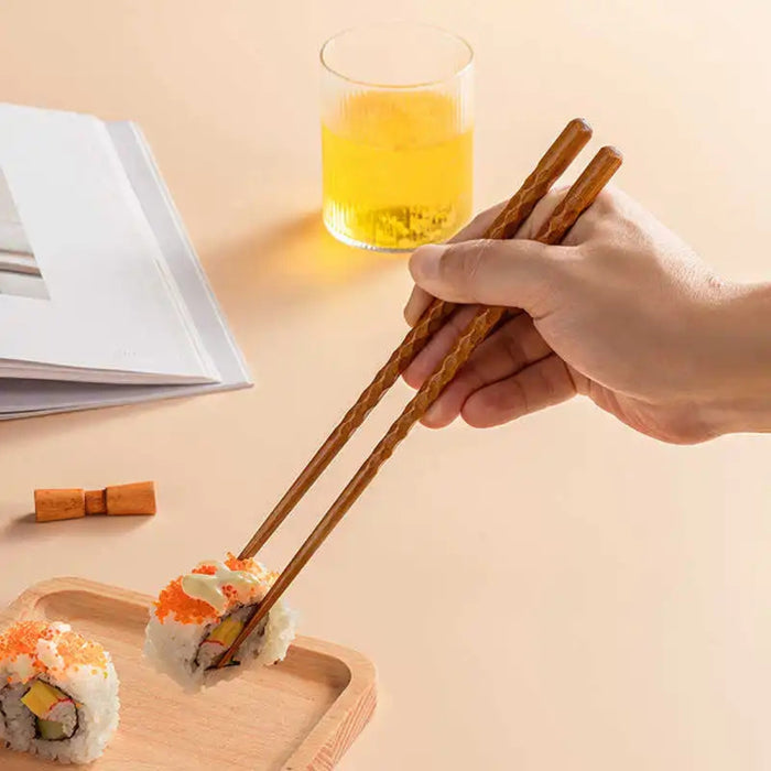 Hand using wooden chopsticks to pick up sushi on a wooden tray with a glass of yellow drink in the background.