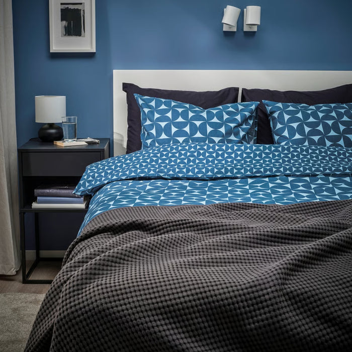 Modern bedroom with IKEA ÄNGSNEJLIKA duvet cover and pillowcases in blue geometric pattern, paired with a dark gray blanket, white headboard, and minimalist decor including wall-mounted reading lights and a black nightstand.

