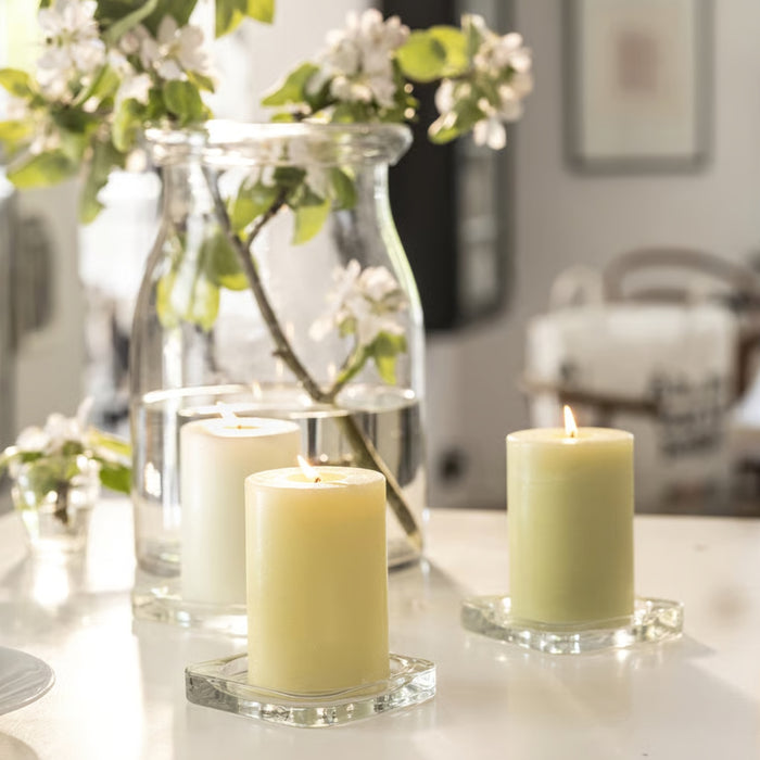 A cozy and elegant home setting featuring two lit IKEA LÖVSKOGSLUND Scented Pillar Candles with apple fragrance, placed on square glass coasters. A large glass jar behind them holds a branch with green leaves and delicate white flowers, creating a refreshing and tranquil ambiance. The soft lighting enhances the warm and inviting aesthetic.-90582065